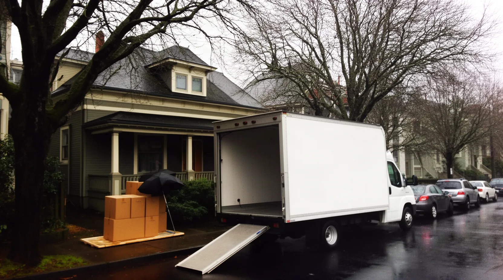 An unmarked box truck parked on a narrow wet eastside Portland street with moving boxes waiting under a bungalow porch